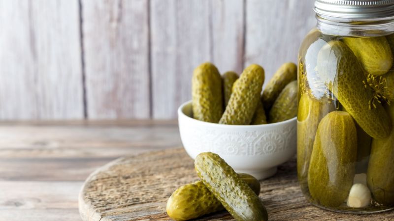 A canning jar full of pickling cucumbers sits on a table. Next to it is a bowl with some of the homemade pickles.
