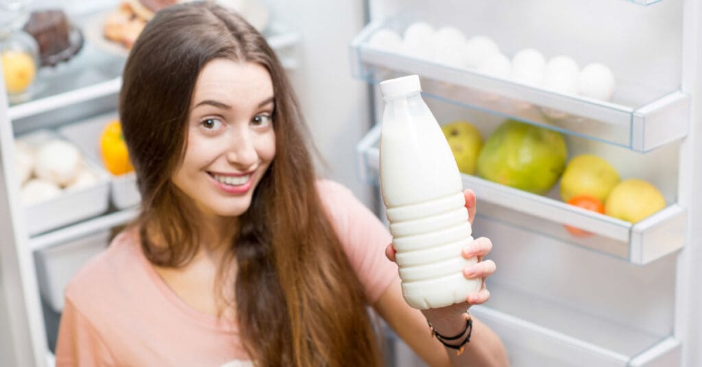A woman holds a plastic container filled with raw milk in front of her fridge. Plastic containers are safe for freezing raw Jersey cow milk