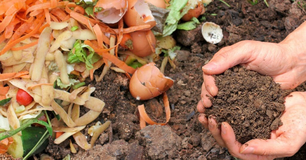 A man holds a pile of compost in his hands next to food scraps like eggshells, potato peels, and other organic materials. He uses them to learn how to compost on his homestead. 