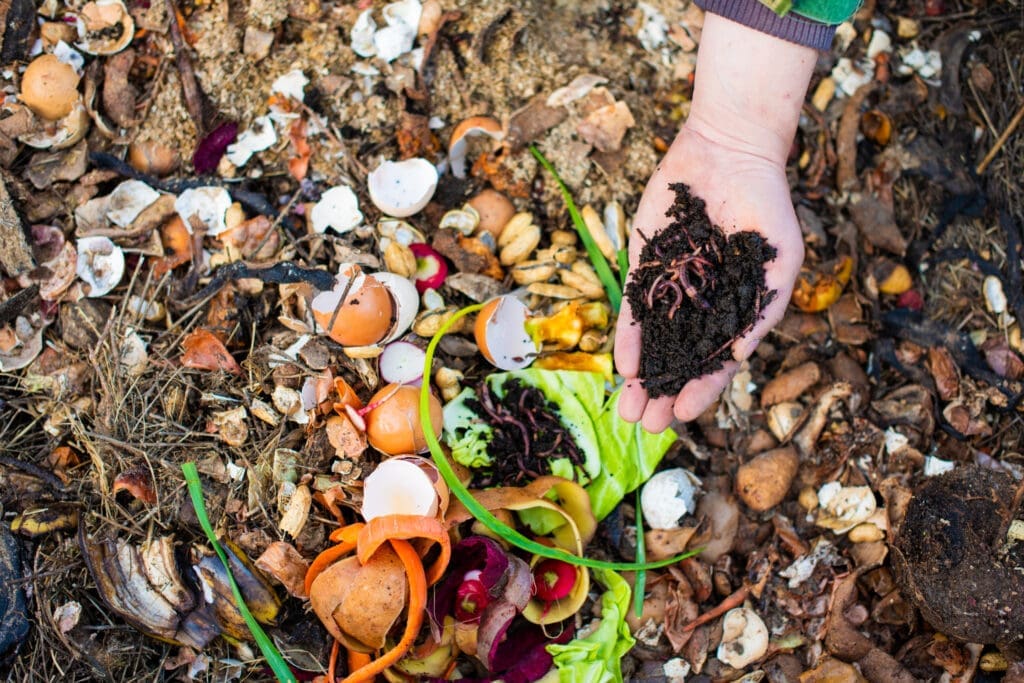 A woman holds a handful of dirt that has worms crawling in it near her compost bin. Learning how to compost requires choosing a method. Vermicomposting is one of the most popular. 
