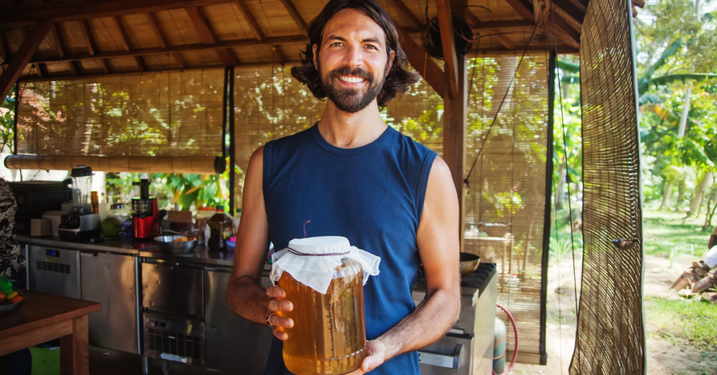 A man holds a gallon-sized glass jar with a cloth top that has homemade kombucha inside. He followed kombucha recipes to make the batch.