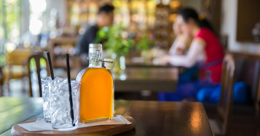A bottle of kombucha and two glasses with ice and straws sit on a table. Nearby is a coiple sitting at another table waiting for the drinks. 